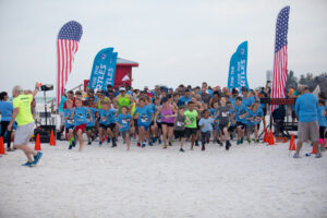 More than 1000 runners, many of them children, sprint across the start line, beginning the race by running directly at the camera. Two American flags stand at either end of the start line and the sidelines are marked with cones. Most wear Run for the Turtles t-shirts.